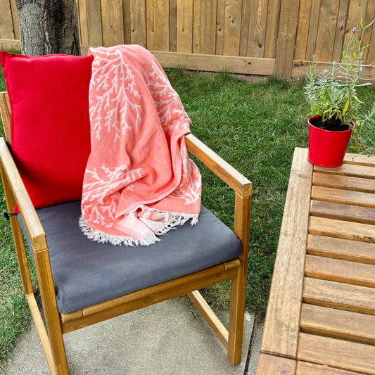 Chair with a red pillow and pink coral Turkish towel throw on a patio with a wooden table and plant in the background.