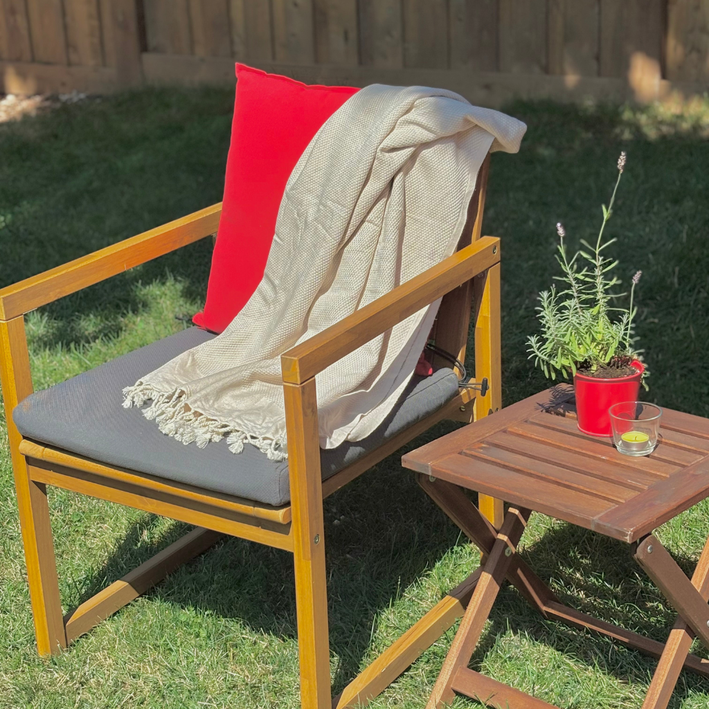 Wooden patio chair with a beige Turkish towel throw and red pillow, accompanied by a small table with a plant and candle on a grassy area.