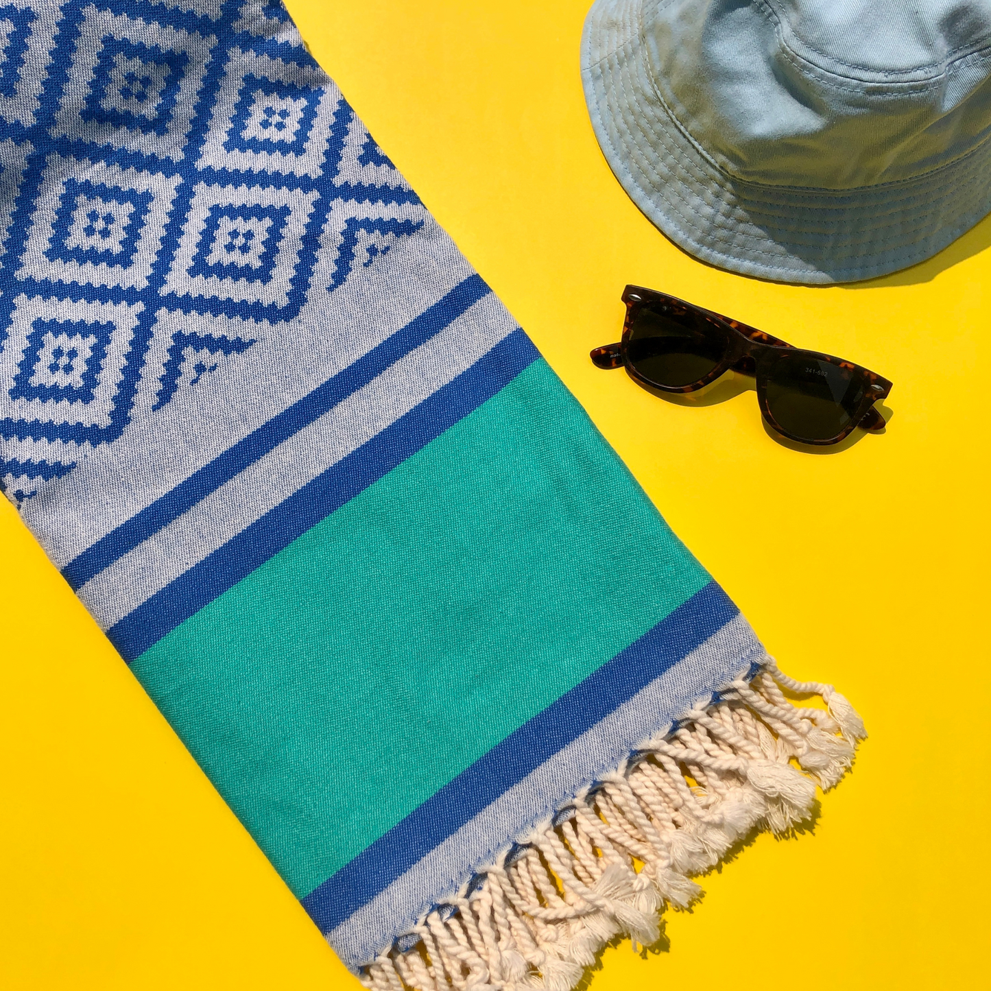 A blue and green Turkish towel with a geometric pattern and tasselled edges is laid out on a bright yellow background besides a pair of sunglasses and a light blue bucket hat.