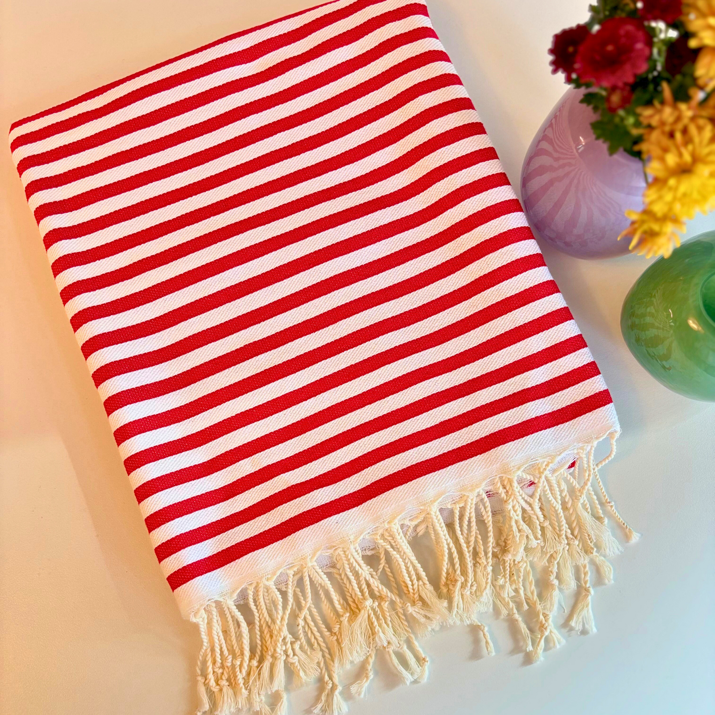 Folded FUN Turkish Towel in wine red with white stripes and hand-knotted fringes, placed next to vases with burgundy and yellow flowers