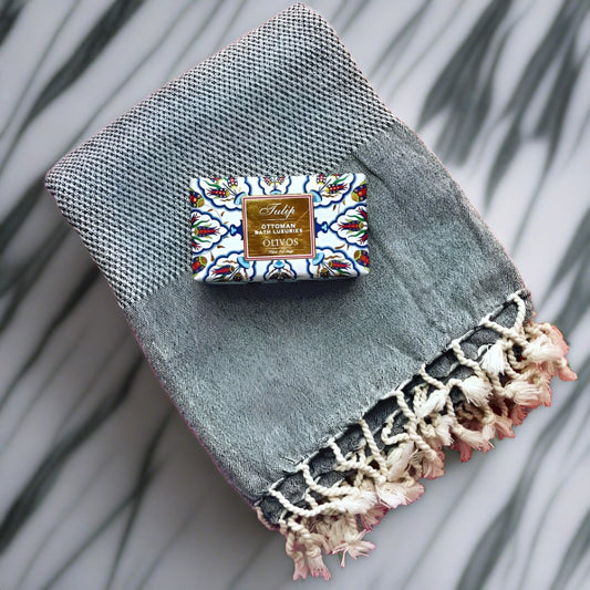 A bar of HAMMAM Olive Oil Soap on a charcoal DENIZLI Turkish bath towel with a marble bathroom background