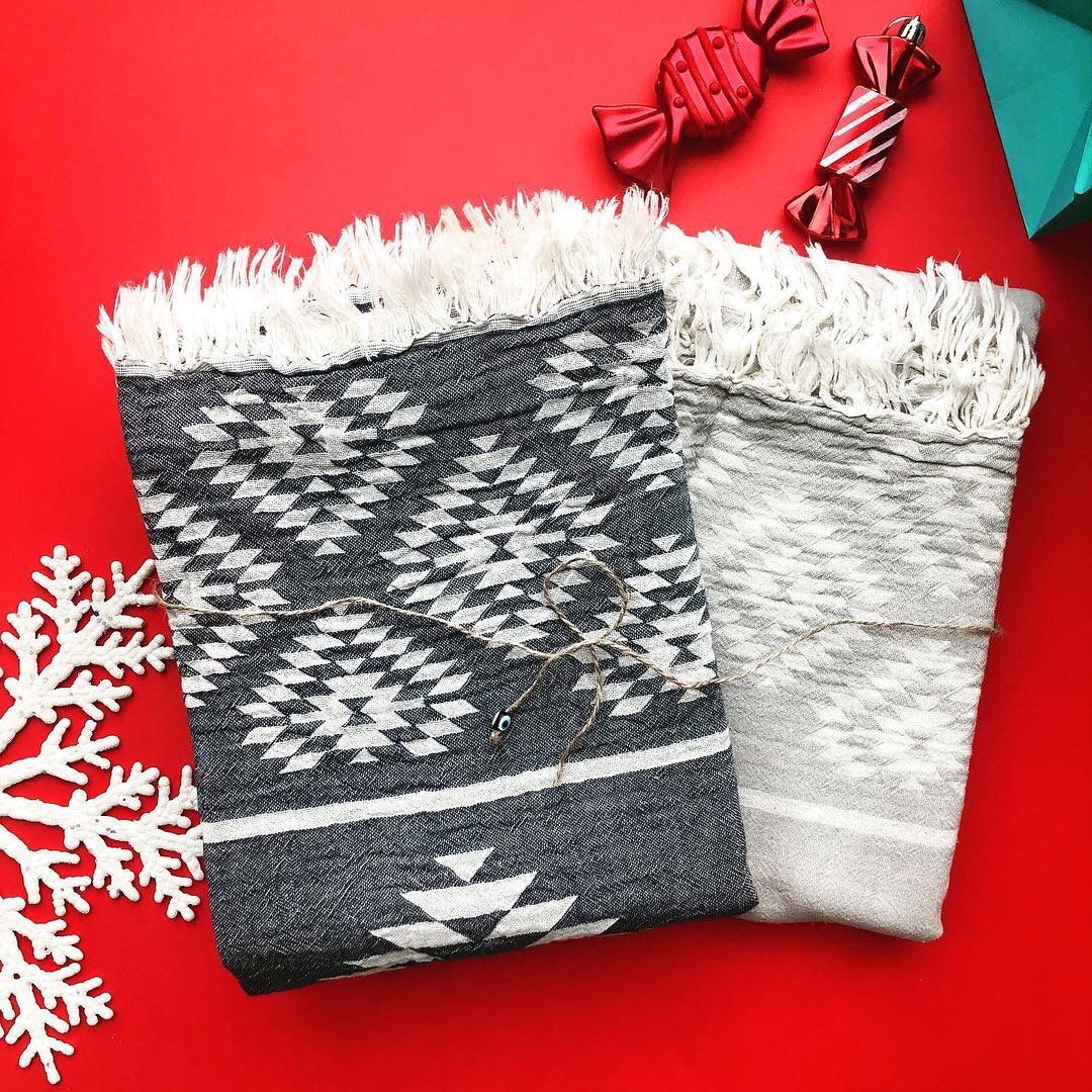 Two reversible Turkish towels with kilim patterns, one in charcoal and white, the other in lighter grey, on a red background with holiday decorations including a white snowflake and candy-striped ornaments