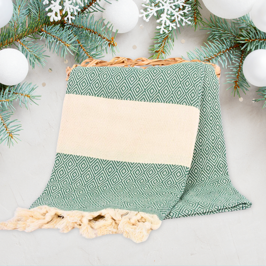 Sage green Turkish towel with a diamond pattern and cream accents, featuring knotted fringes, displayed on a festive holiday background with evergreen branches and snowflakes, ideal for seasonal home decor or gifting