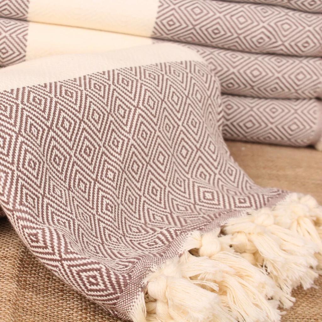 MOCHA Pera Turkish Towel in a warm brown-beige colour with diamond weave pattern and hand-tied fringes, displayed rolled on a textured fabric surface.