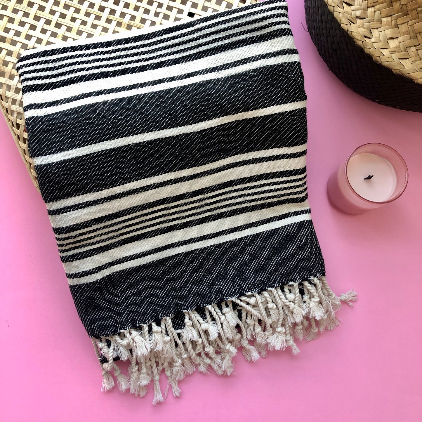TOPKAPI Turkish Towel with black stripes, folded and displayed on a rattan box, beside a striped rattan basket and pink candle on a pink background.