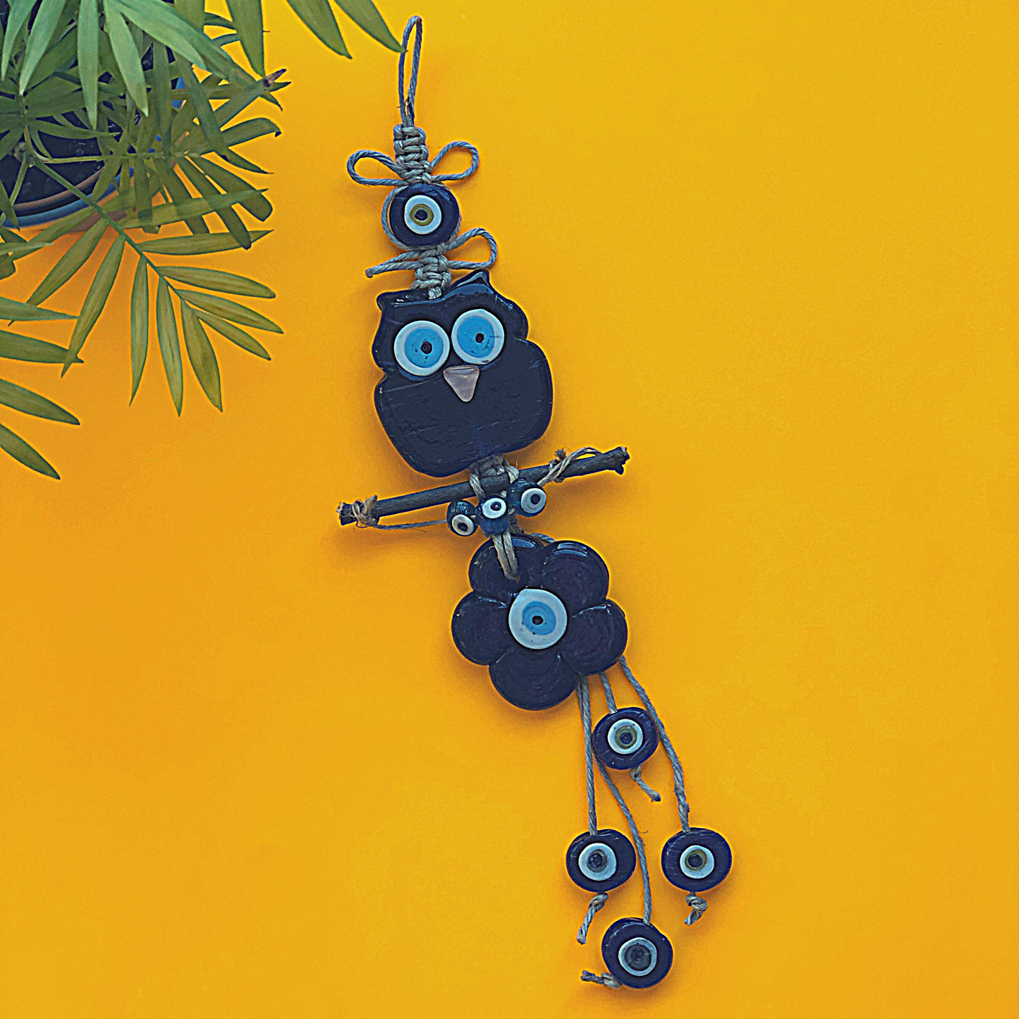 Handmade Turkish evil eye wall hanging featuring a glass owl and a daisy, crafted with jute rope, displayed on a bright yellow background with green leaves.