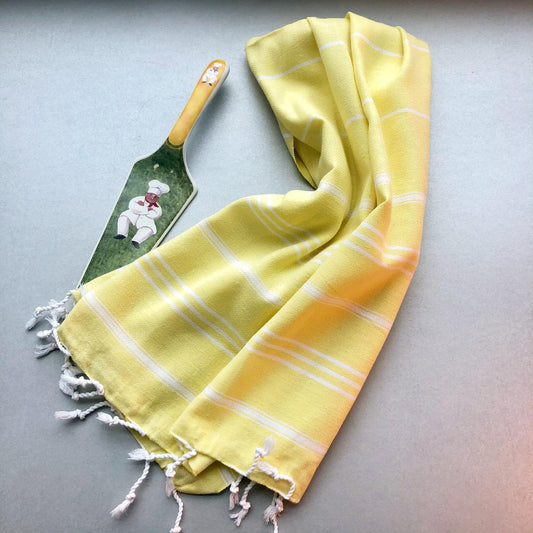 A yellow STRIPY Turkish kitchen towel in the 'Lemon' colour variant, featuring white stripes and fringed edges, is draped on a light surface next to a decorative spatula with a chef illustration.