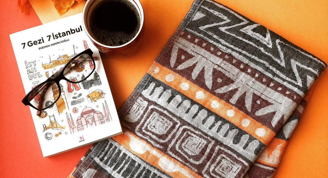 A cozy autumn setting on an orange background featuring a book about Istanbul, reading glasses, a cup of warm coffee, and a folded Turkish towel in warm fall colours