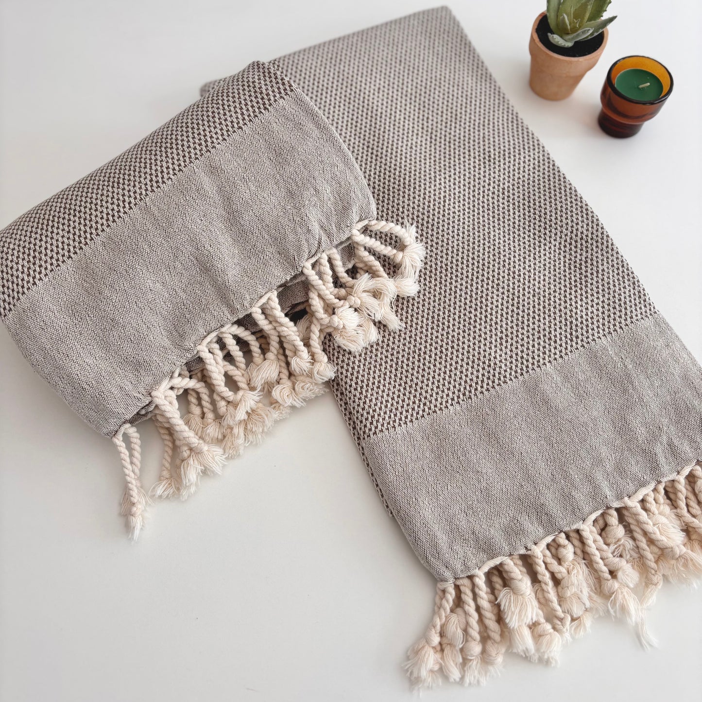 Light brown cotton Turkish towel with tassels on a white surface next to plant and tealight candle.