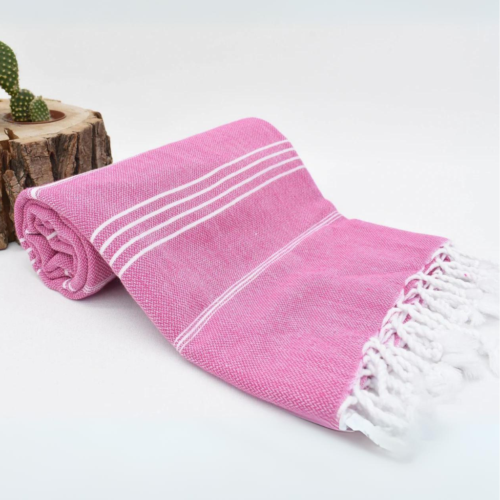 Pink Turkish towel with white stripes & tassels, rolled up on a white surface next to a cactus, ideal for beach, bath or travel use.
