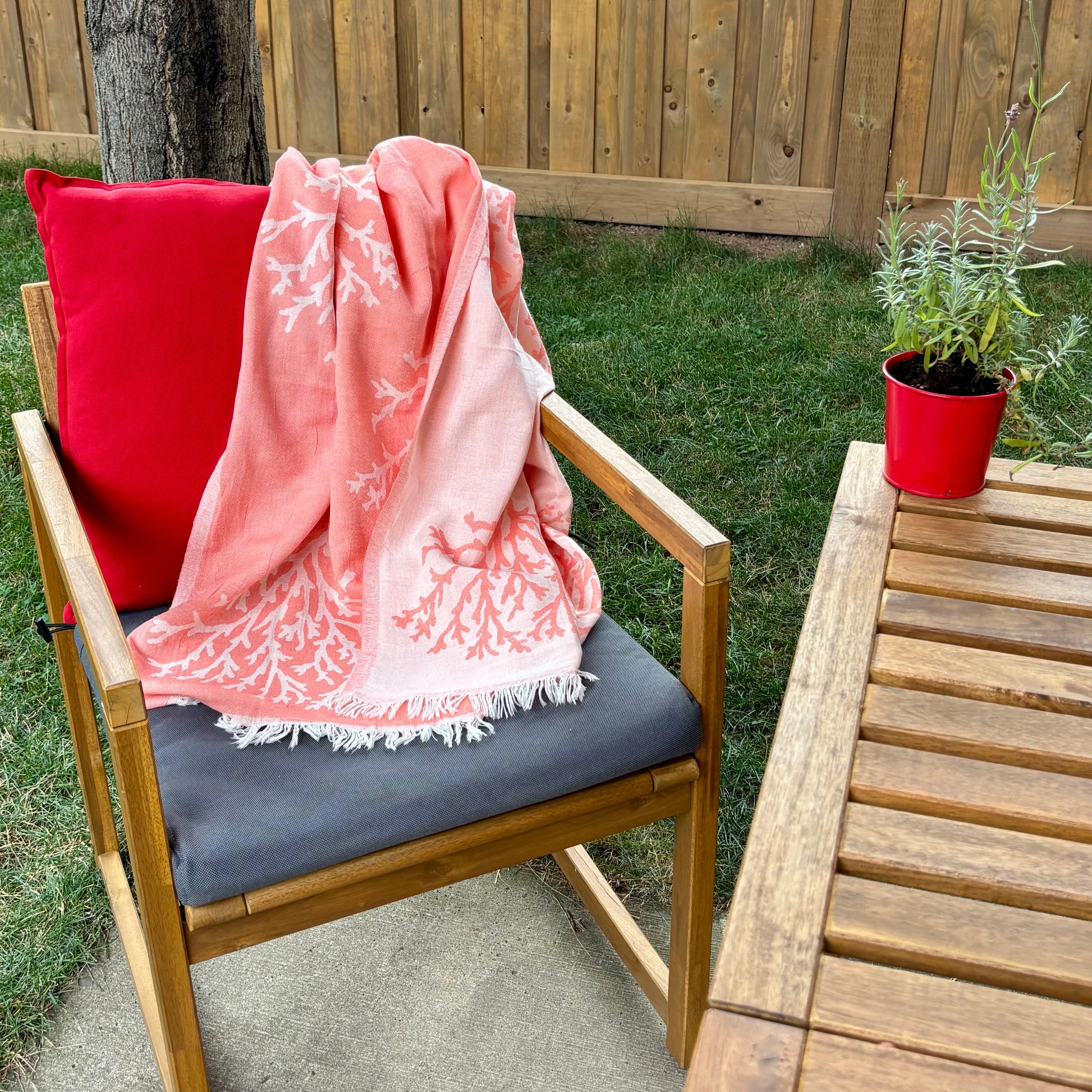 Chair with a pink coral reversible Turkish towel and red pillow on a patio with a wooden table and potted lavender.