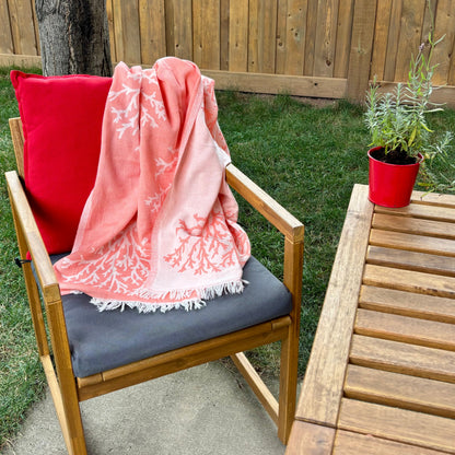 Chair with a pink coral reversible Turkish towel and red pillow on a patio with a wooden table and potted lavender.