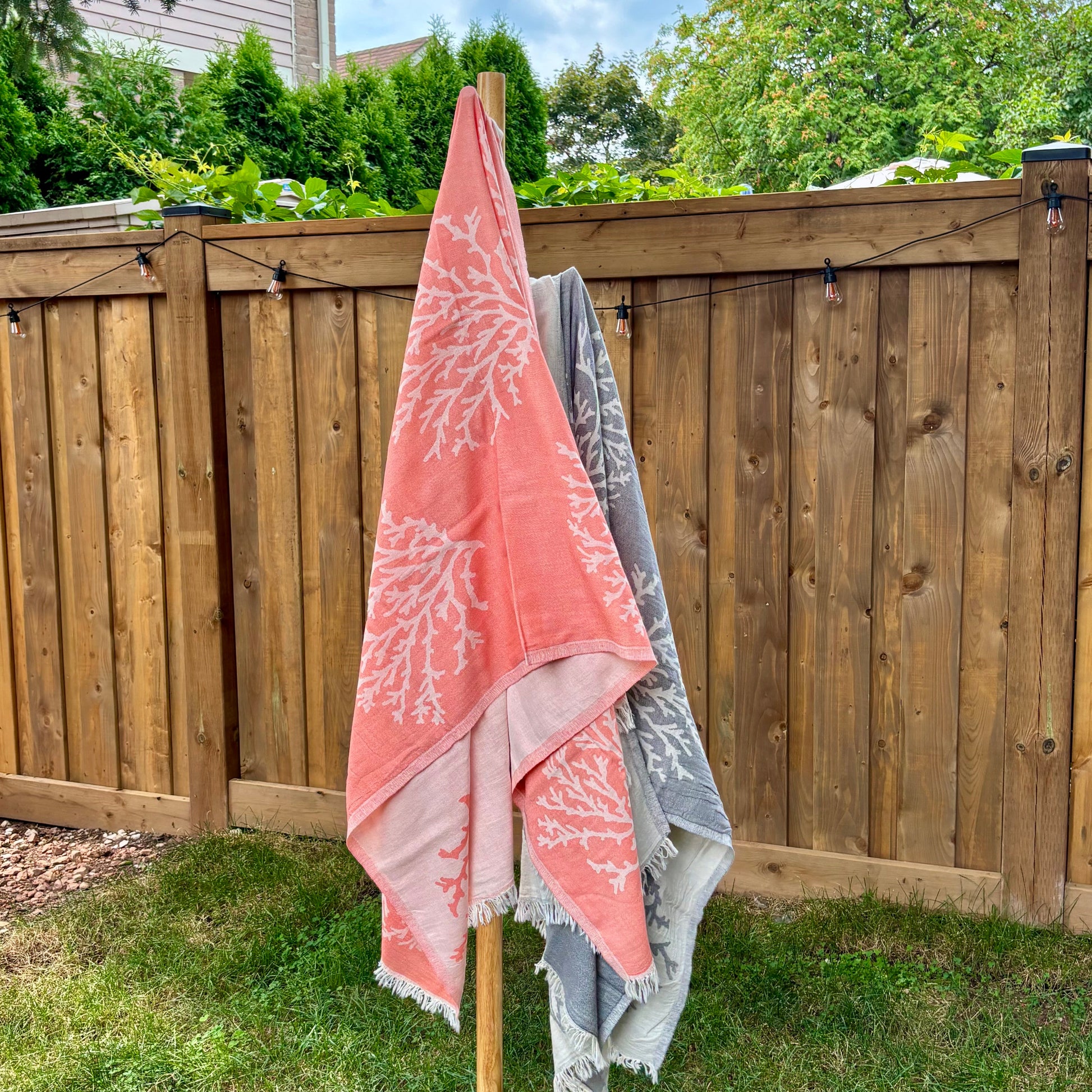 Two coral-patterned Turkish towels, one pink and one grey, hanging on a wooden stand outdoors.
