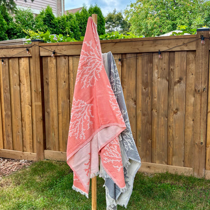 Two coral-patterned Turkish towels, one pink and one grey, hanging on a wooden stand outdoors.