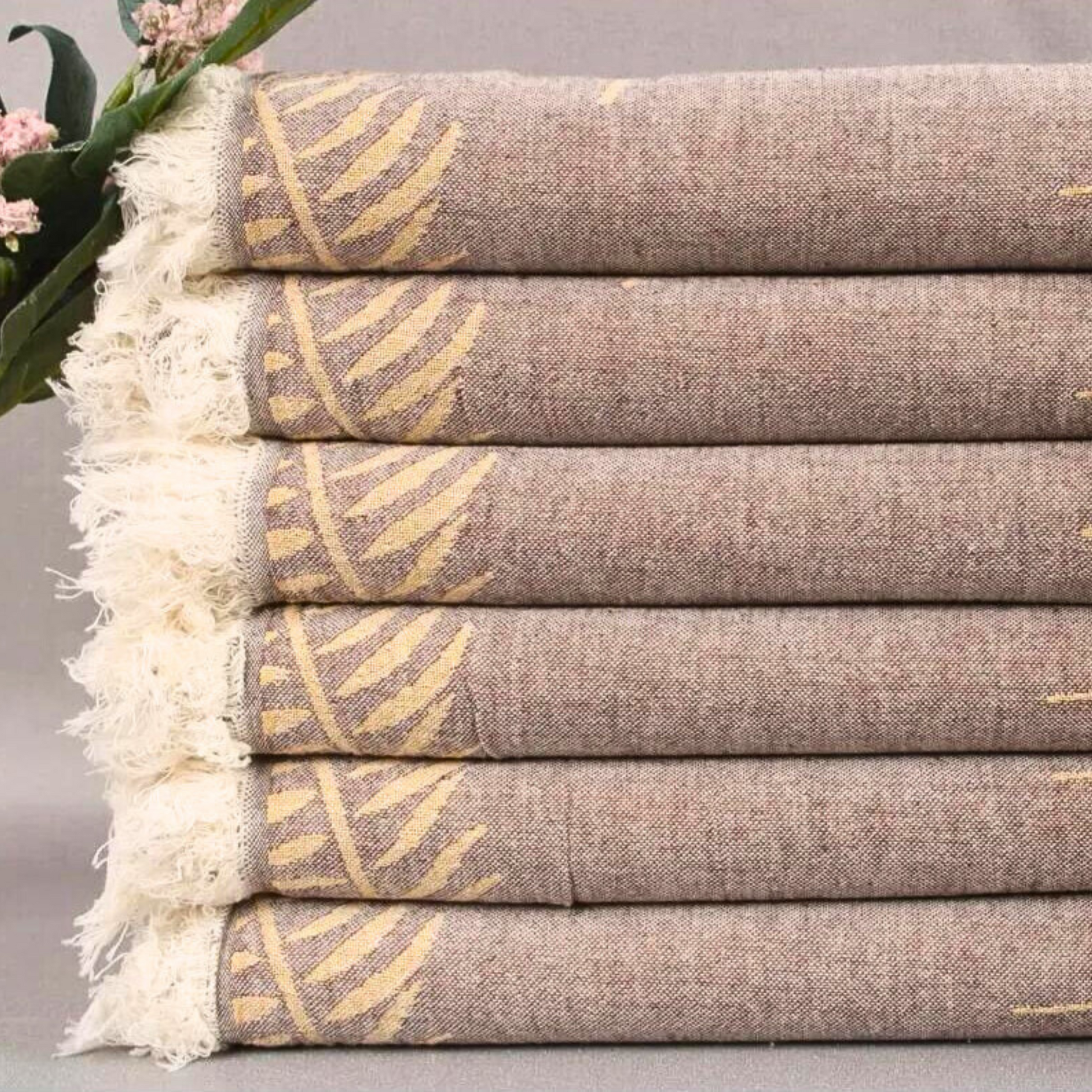 Stacked brown FERN Turkish Towels with yellow leaf patterns, with a white fringed edge, set against a backdrop of soft pink flowers and a grey background
