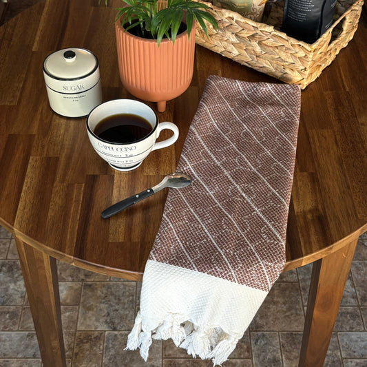 Folded brown WALNUT Turkish Hand Towel on wooden table with a cup of coffee, plant, and coffee basket.