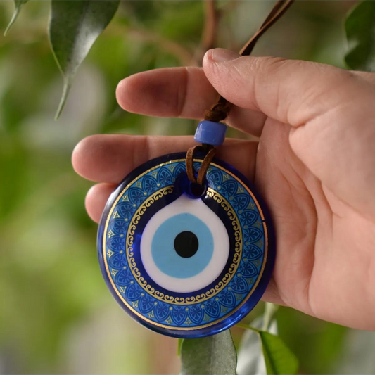 A hand holding a Nazar wall hanging with intricate blue patterns, suspended against a backdrop of green leaves