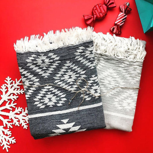 Two reversible Turkish towels with kilim patterns, one in charcoal and white, the other in lighter grey, on a red background with holiday decorations including a white snowflake and candy-striped ornaments