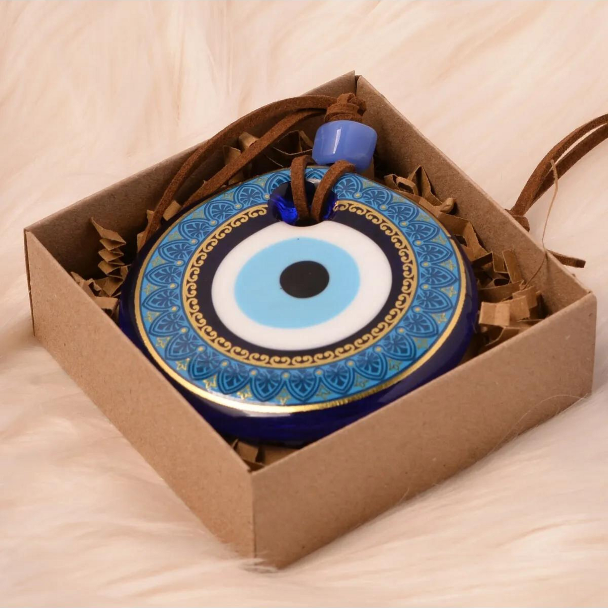 A Nazar wall hanging, adorned with blue and gold details, resting inside a cardboard gift box on a soft, white surface