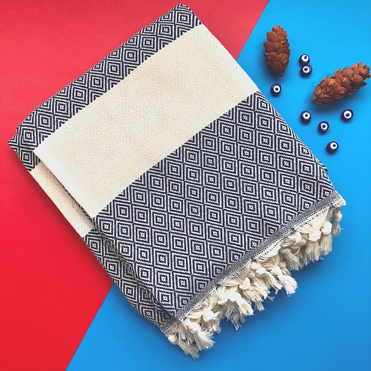 PERA Turkish Towel in navy, featuring geometric patterns and a beige stripe in the center, neatly folded on a festive background with pine cones and evil eye beads scattered nearby