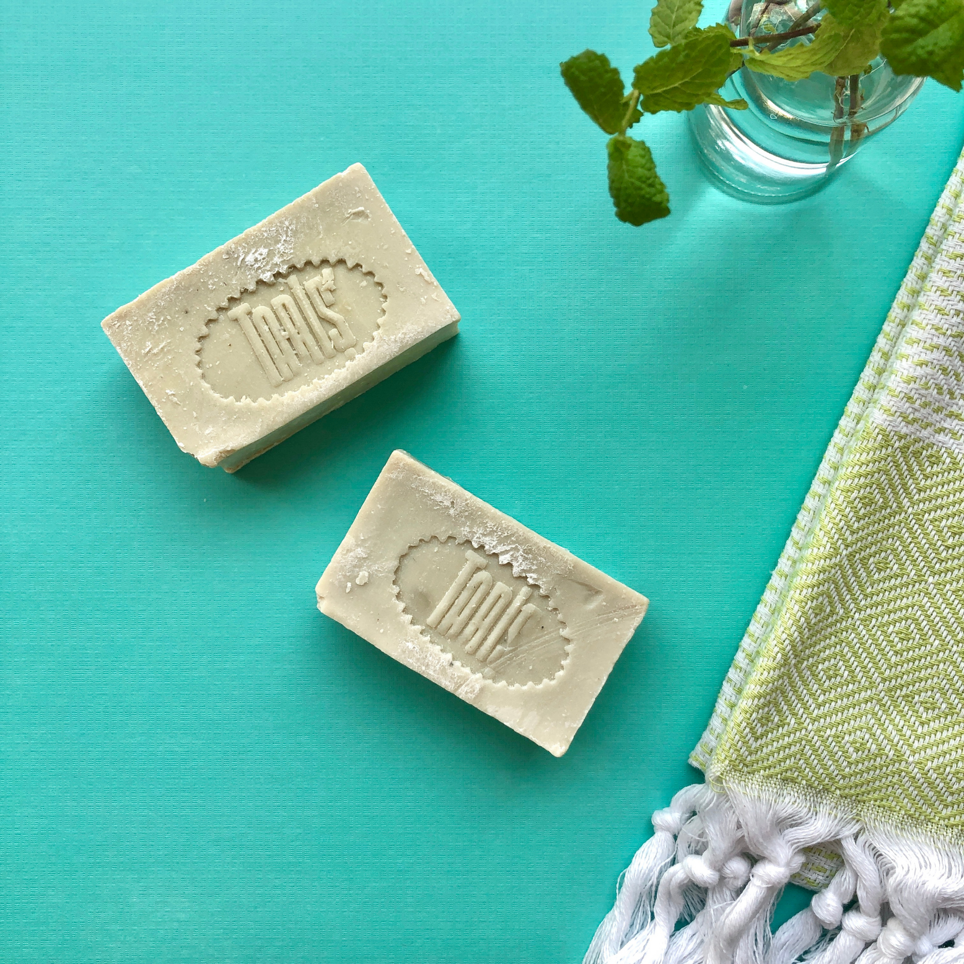 2 bars of OLIVE OIL Soaps next to green Turkish hand towel and mint branches in a vase on turquoise background