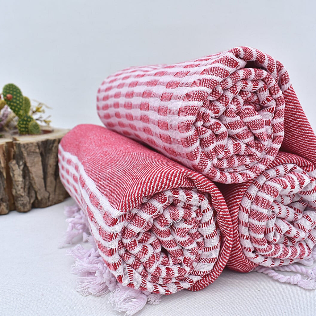 Rolled red and white checkered Turkish towels with cactus and wooden planter decoration on a white backdrop