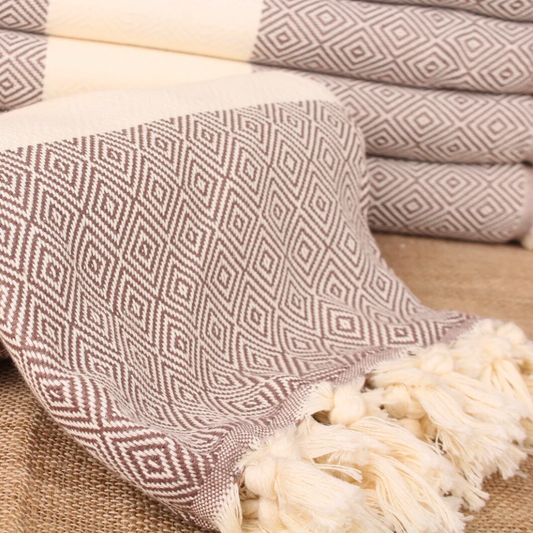 MOCHA Pera Turkish Towel in a warm brown-beige colour with diamond weave pattern and hand-tied fringes, displayed rolled on a textured fabric surface.