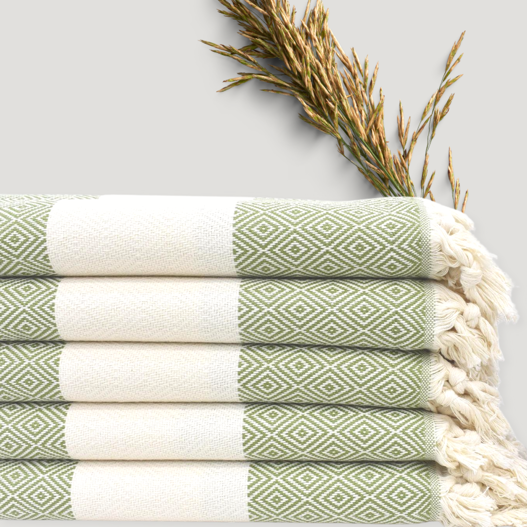 PERA Turkish Towels in sage, stacked neatly with intricate geometric patterns visible, accompanied by dried golden plants on a light backdrop