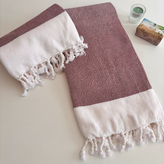 RUBY Turkish Towel featuring a burgundy and white diagonal twill pattern with hand-tied fringe, styled on a white table with artisanal soap.