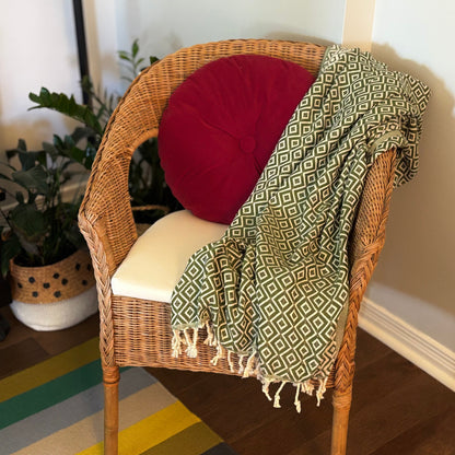 Wicker chair with a patterned green and white THYME Peshtemal Towel draped over it, next to a round dark red pillow, on a wooden floor with a colourful striped rug.